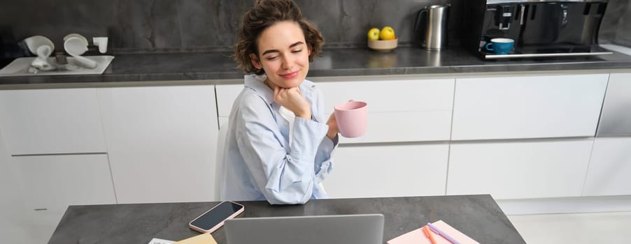 Beautiful woman managing her work from home, drinking coffee and looking at laptop, sitting in kitchen with paperwork, doing her taxes, studying indoors on remote.