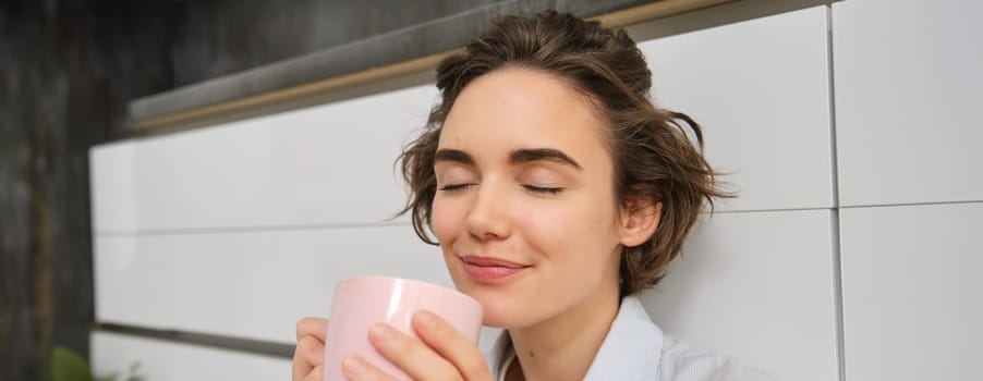 Coziness and home concept. Young woman enjoys her morning cup of coffee, drinks fresh tea and smiles from comfort and relaxation, sits on kitchen floor with pink mug.