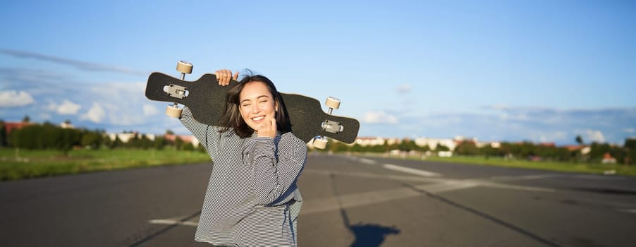 Leisure and people. Happy asian woman standing with longboard, cruising on an empty road in countryside. Skater girl holds her skateboard and smiles at camera.