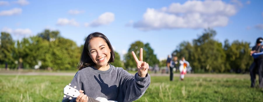 Vertical shot of positive asian girl shows peace sign, plays ukulele in park, rests and enjoys the day. Copy space.