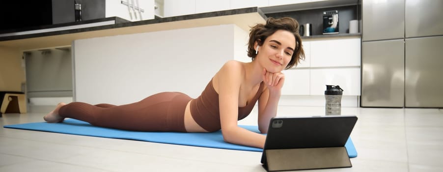 Portrait of smiling girl in fitness tracksuit, lying on mat at home, looks at tablet, watches online sports video tutorial, meditaiton lesson, yoga or workout online gym instructor.