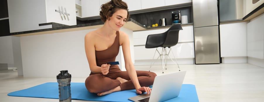 Portrait of young female athlete, yoga girl paying for online classes, purchasing remote training with gym instructor, sitting on her rubber mat with laptop and credit card.