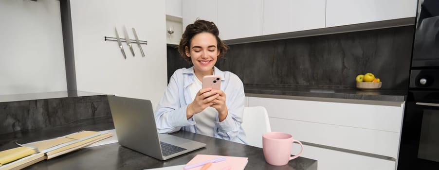 Portrait of brunette woman chats on mobile phone, uses smartphone app from home, orders delivery while sits in kitchen.