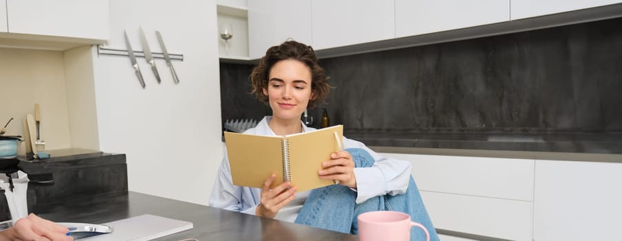 Young woman sitting in kitchen, reading her notes in notebook. Smiling girl at home, doing homework, studying.