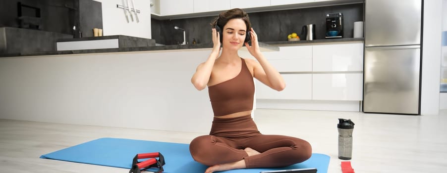 Smiling fitness girl in headphones, watches pilates, gym instructor online video, workout at home on yoga rubber mat, uses plank equipment and resistance band.