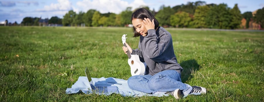 Smiling asian girl learns how to play ukulele via laptop, online video tutorials, sitting on grass in park with musical instrument.