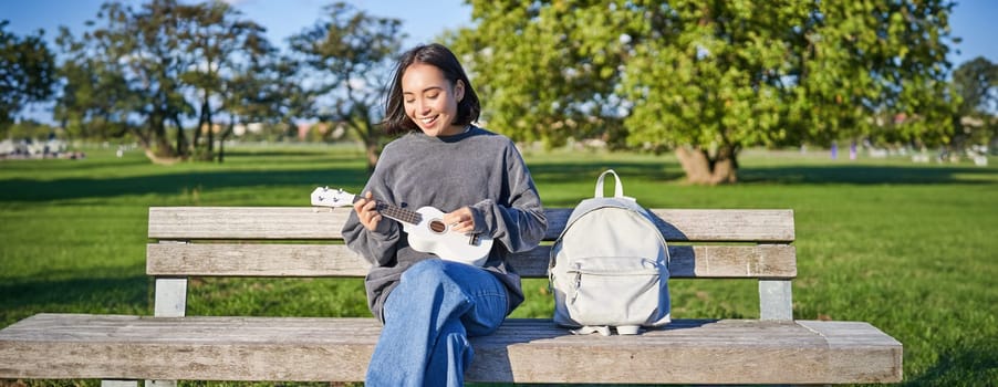 Beautiful asian girl plays ukulele outdoors, sits in park on bench with musical instrument.