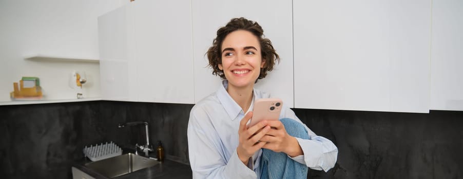 Smiling brunette girl sitting with smartphone at home, orders takeaway from mobile phone app, using online shopping application.