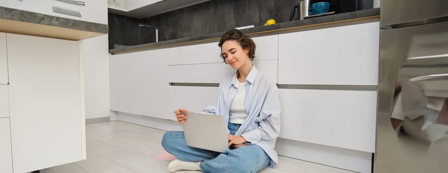 Freelance and lifestyle concept. Young woman sits on floor with laptop, works from home, sets up her remote workspace in kitchen, does homework.