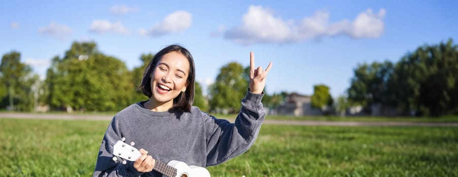 Happy asian girl playing ukulele in park, showing rock n roll, heavy metal horns sign and smiling, having fun outdoors.