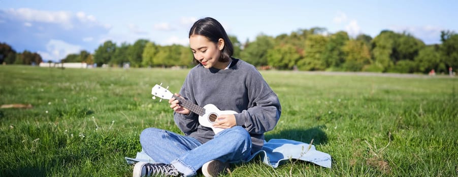 Beautiful asian girl sitting in park, playing ukulele and singing, relaxing outdoors on sunny spring day.