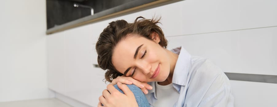 Close up portrait of beautiful, tender young woman smiling, looking at camera, sitting in cozy pose on kitchen floor, daydreaming.