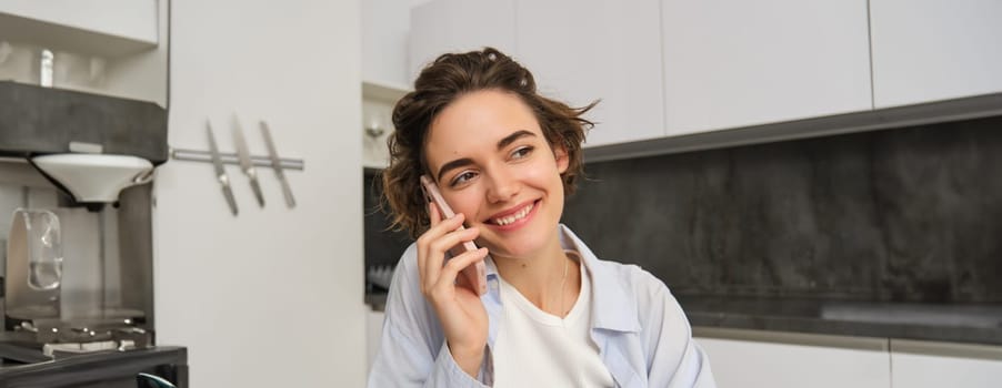 Portrait of young woman, 25 years old, holding smartphone, talking on mobile phone, sitting in kitchen at home.