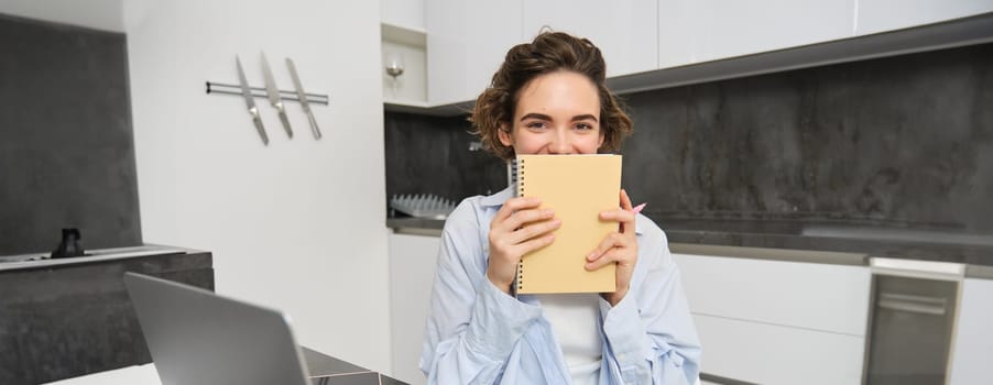 Portrait of smiling brunette girl, holds her planner, writes in notebook and looks happy, laughs, does homework, makes notes, sits at home in kitchen.