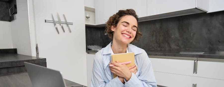 Portrait of young creative woman, holding her notebook and smiling, makes notes in her diary, writing in her planner, sitting at home in kitchen and smiling.