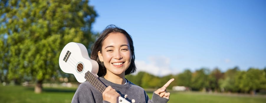 Portrait of asian smiling girl, holding ukulele over shoulder, pointing finger at copy space, banner or logo.