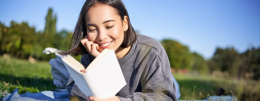 Portrait of beautiful smiling asian girl, reading in park, lying on grass with favourite book. Leisure and people concept.