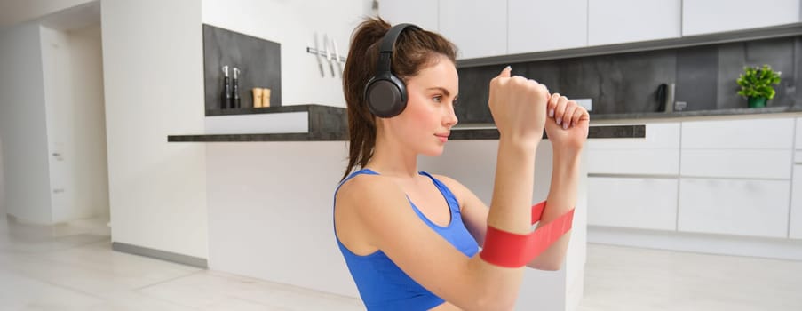 Portrait of determined beautiful woman, listening music, workout with elastic resistance band, doing exercises on arms, wearing sportsbra.