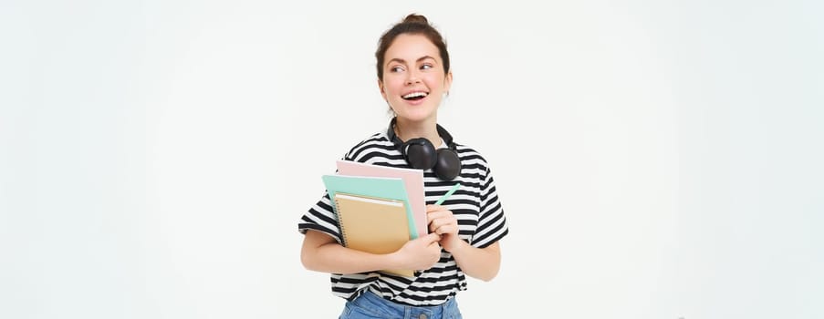 Student and education concept. Young woman with books, notes and pen standing over white background, college girl with headphones over neck posing in studio.