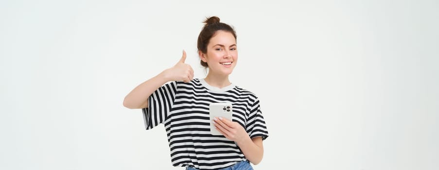 Cheerful, smiling young woman shows thumbs up, approves and recommends mobile app, holds smartphone, looks satisfied, stands over white background.