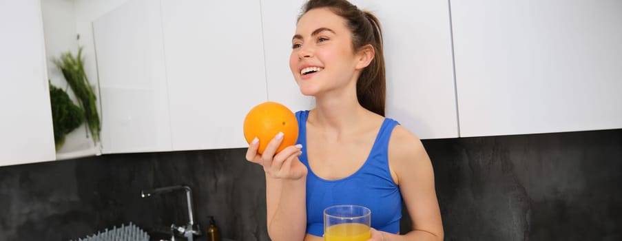 Indoor shot of woman after workout, standing in kitchen with fresh juice and an orange, drinking it.