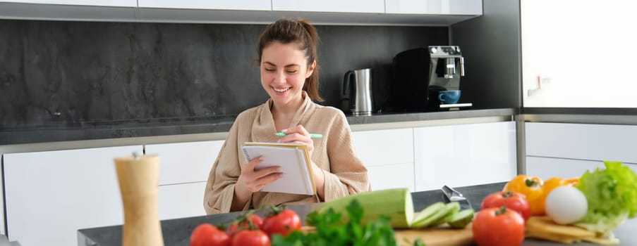 Portrait of beautiful, smiling young woman making list of meals, writing down recipe, sitting in the kitchen with vegetables, doing house errands.