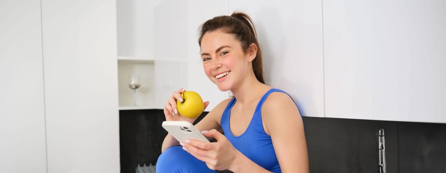 Portrait of brunette fitness woman, eating an apple, holding smartphone, using mobile phone app while having healthy fruit snack in kitchen.