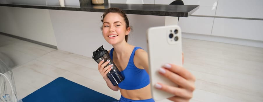 Portrait of woman doing sports, takes selfie on smartphone, fitness instructor records her exercises, stays hydrated, drinks water during training session.