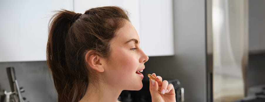 Close up portrait of fitness girl, taking vitamins, holding pill dietary supplement, swallowing tablet buds for healthy and strong body, standing in kitchen.