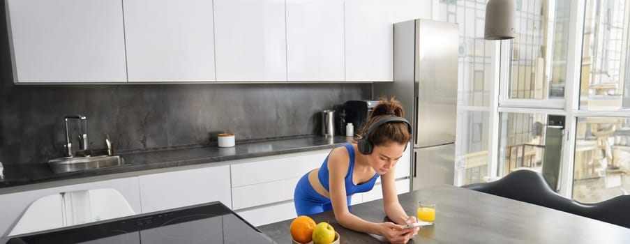 Portrait of brunette woman in sportswear, using smartphone, standing in kitchen, drinking fresh orange juice, watching workout gym tutorials.