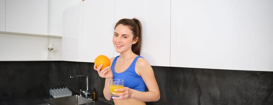 Wellbeing and sport. Young smiling nutritionist, fitness girl holding orange and fresh juice, drinking it from glass and looking happy, posing in sportsbra and leggings, standing in kitchen.