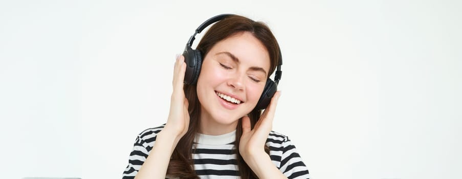 Portrait of beautiful woman in wireless headphones, listening music, using earphones, smiling at camera, standing over white background.