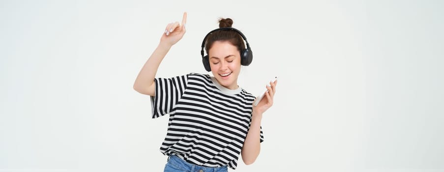 Portrait of happy brunette woman, dancing in headphones, listening music on her mobile app, enjoys her favorite song, isolated against white background.