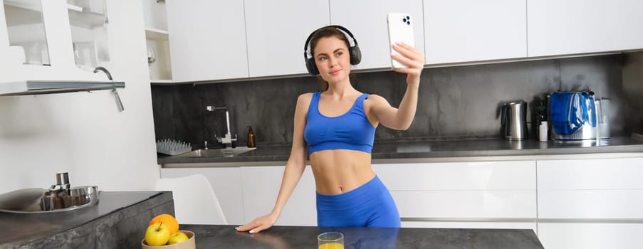 Portrait of carefree sportswoman, dancing in kitchen with smartphone, listening music in headphones, feeling energized and excited after workout.
