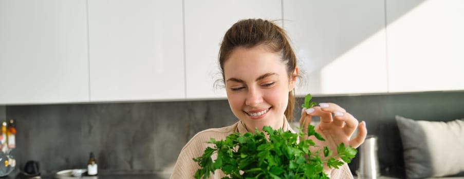 Portrait of attractive girl with bunch of parsley, eating fresh herbs and vegetables, cooking in the kitchen.