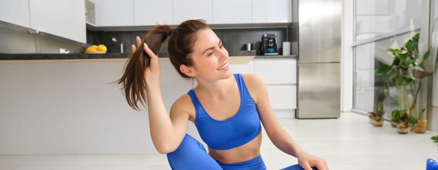 Portrait of young, healthy and fit brunette woman, sits on rubber mat, does yoga, workout training at home, wears blue fitness sportswear and smiles.