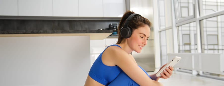 Portrait of brunette girl in fitness clothes, listens music in wireless headphones, holds smartphone, workout from home, looking at training video instructions.