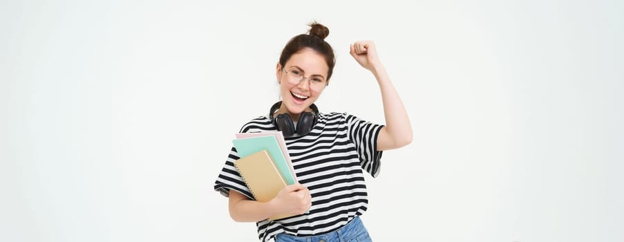 Portrait of young woman in glasses, wearing headphones over neck, holding planner, notebooks and study material, cheering, rooting for you, celebrating, standing over white background.