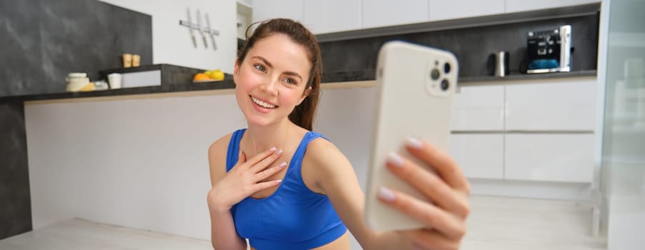 Smiling beautiful girl records video while doing sports at home, looks at smartphone, takes selfie on mobile phone, workout indoors in blue leggings and sportsbra.