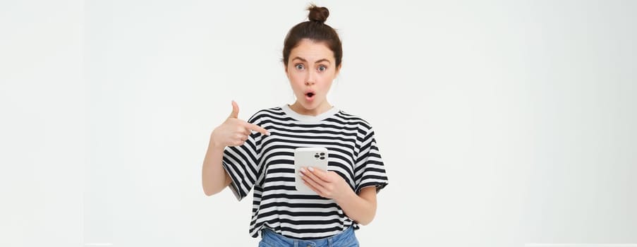 Portrait of impressed woman, drops jaw, points at her smartphone, sees amazing news online on mobile phone, isolated over white background.