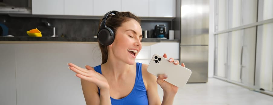 Close up portrait of young happy woman, workout at home, listens music in wireless headphones, singing at smartphone, doing training exercises.