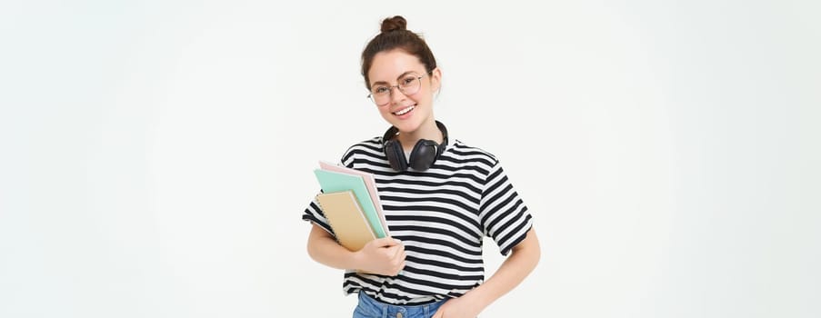 Young woman with notebooks, books and study material, posing over white background, wears headphones over neck.