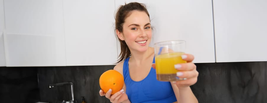 Portrait of smiling, beautiful young woman, offering glass of fresh juice, holding an orange and looking happy, fitness instructor giving you a drink.