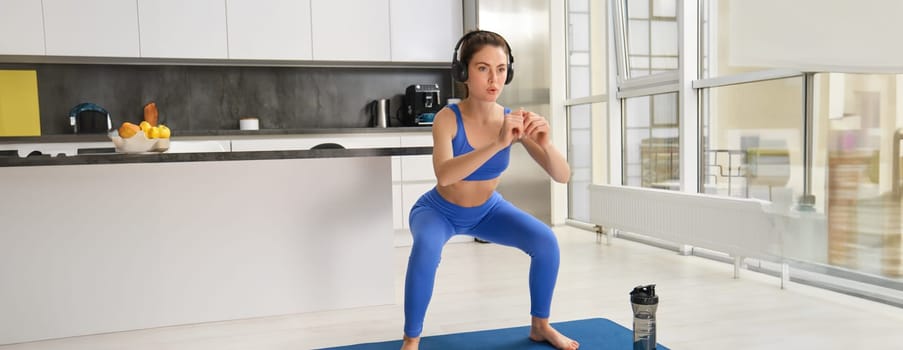 Portrait of young woman doing aerobics exercises in living room, sport training at home, standing on rubber fitness mat and doing workout.