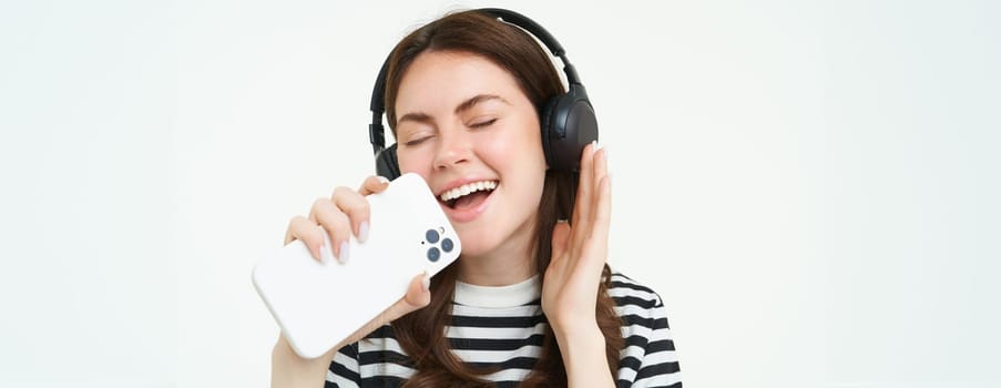 Portrait of happy girl playing karaoke app, singing into smartphone microphone, wearing wireless headphones, white background.