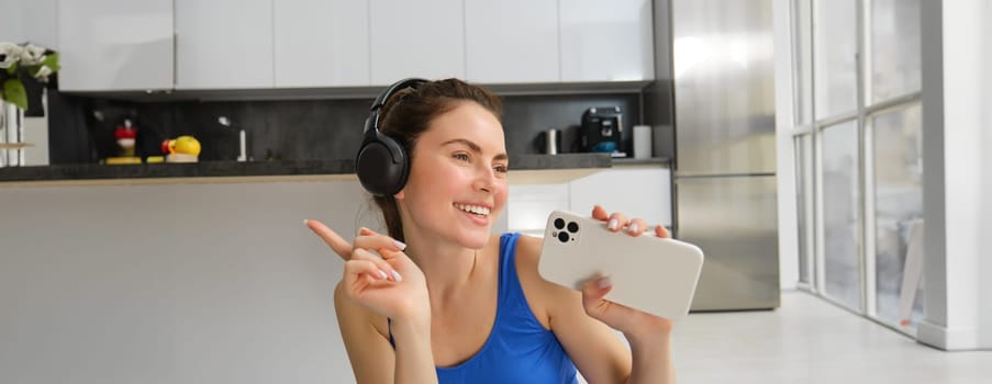 Close up portrait of young happy woman, workout at home, listens music in wireless headphones, singing at smartphone, doing training exercises.