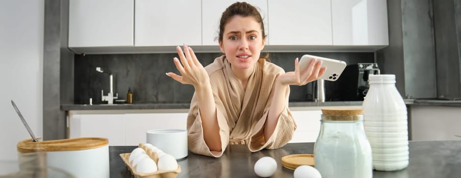 Attractive young cheerful girl baking at the kitchen, making dough, holding recipe book, having ideas.