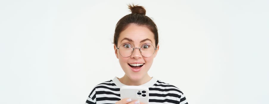 Image of young woman in glasses, using her mobile phone, standing with smartphone and smiling, standing over white background.