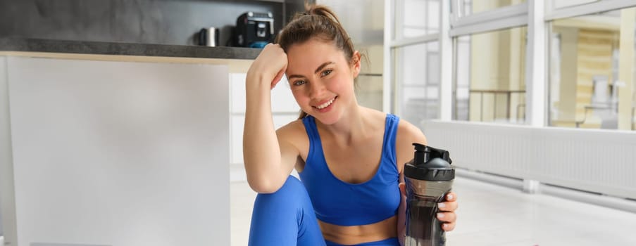 Smiling fitness girl drinks water in the middle of training session, smiling and looking happy, workout from home in blue sportsbra.