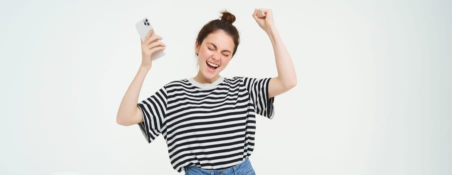 Girl dancing with mobile phone against white background. Woman with smartphone chanting, raising hands up and celebrating.